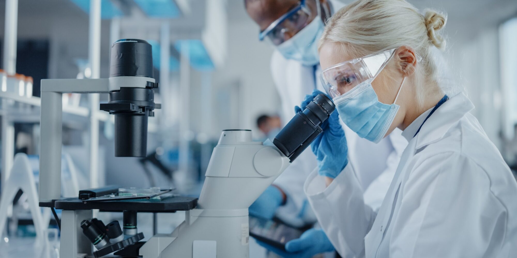 Modern Medical Research Laboratory: Two Scientists Wearing Face Masks Working Together Using Microscope, Analysing Samples, Talking. Advanced Scientific Lab for Medicine, Biotechnology.