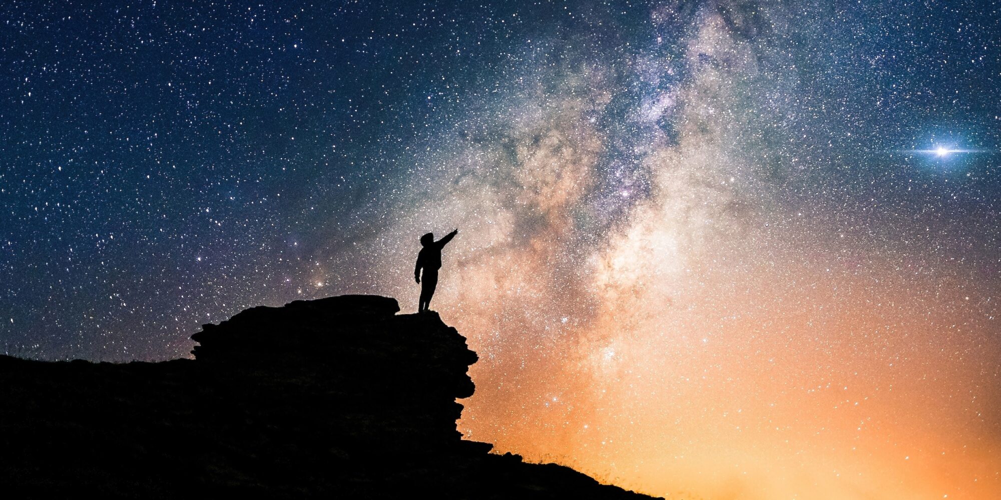 Lone person standing on top of rock, pointing with hand towards the Milky Way and the vast expanse of universe. Stunning silhouette of man gazing out into the cosmos. Power of dreams and aspirations.